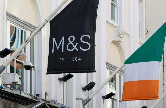 Marks and Spencer shop store logo signage beside Irish tricolor national flag. UK retailer in Dublin, Ireland, Europe with Brexit Covid supply chain issues